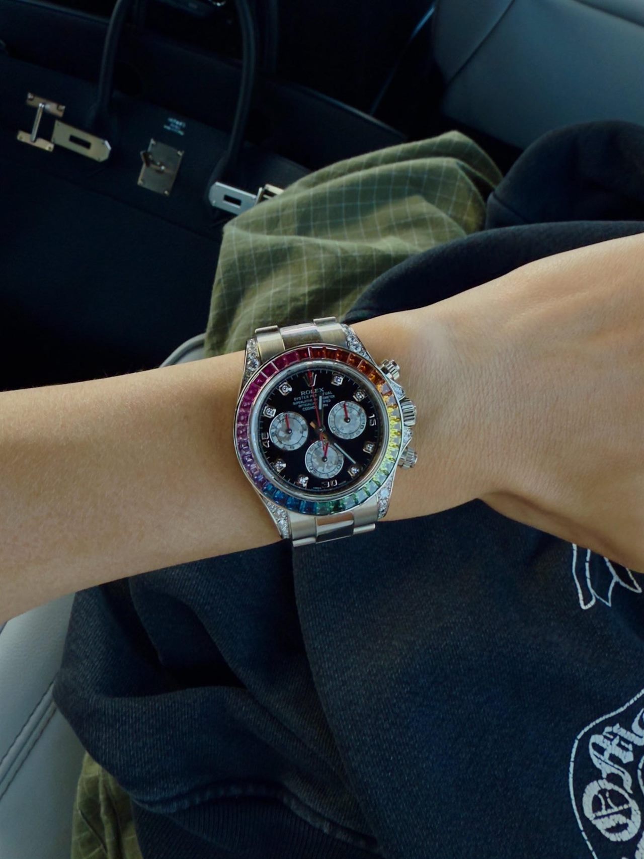 A rainbow-bezel chronograph on a wrist in a car, a black bag beside it
