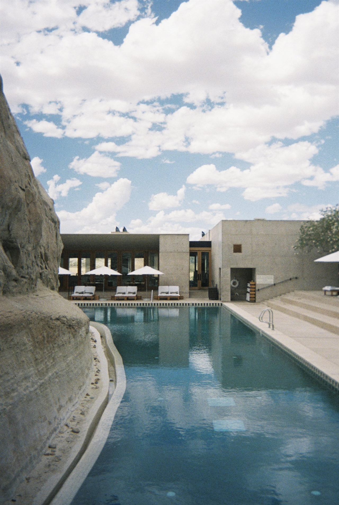 A long pool cut beside a rock face, white umbrellas under a concrete pavilion