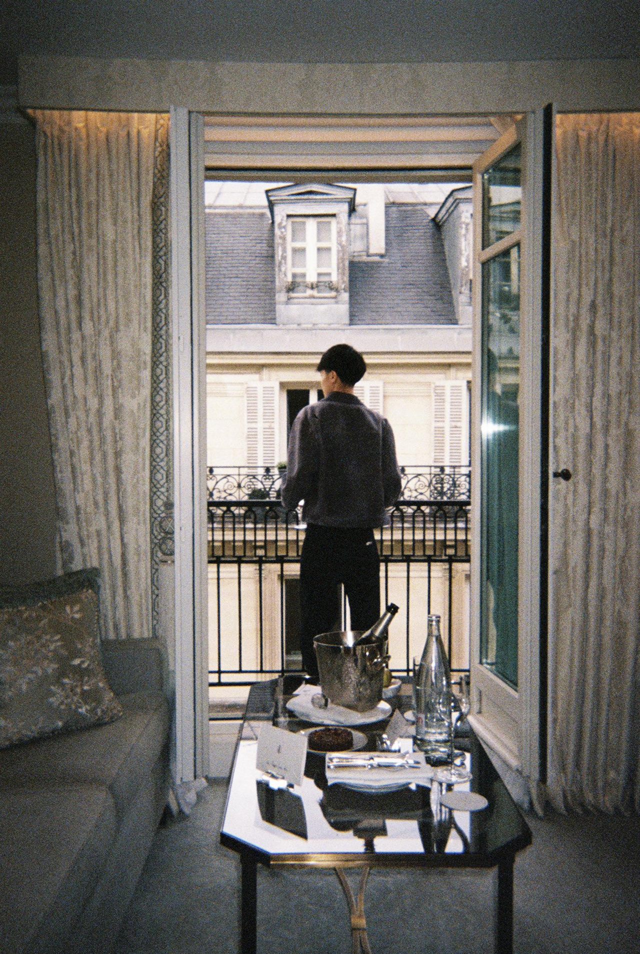 A figure on a Paris balcony seen through open French doors, breakfast on a glass table
